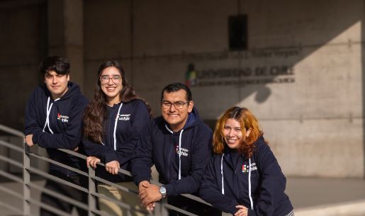 Cuatro estudiantes, dos mujeres y dos hombres, con el polerón de admisión uchile, posando para la cámara, apoyados en una baranda.