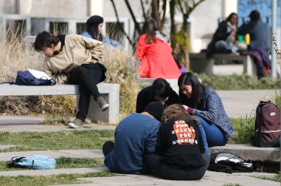 Se pueden ver estudiantes sentadas y sentados, conversando y estudiando, en el patio del Campus Juan Gómez Millas de la Universidad de Chile.