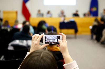 Se ve en primer plano las manos de una persona sosteniendo un celular para sacar una foto. De fondo, borroso, se ve la sala Eloísa Díaz llena de gente y la testera con charlistas.