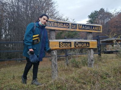 En el exterior, con pasto y árboles de fondo, el licenciado Nicolás Silva, posa frente a señalética de madera que dice "Universidad de Magallanes, sede Omora"