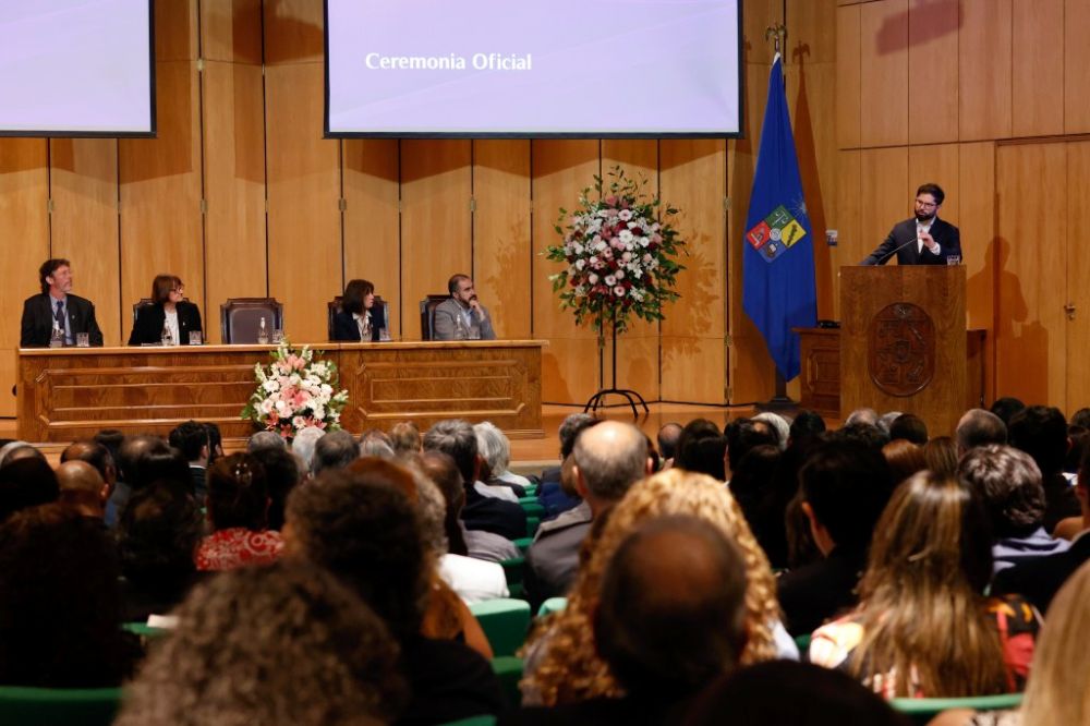 En la imagen se puede ver el Aula Magna de la Facultad de Economía y Negocios, desde el primer piso, con cada una de las butacas ocupadas y al fondo podemos ver, en el estrado dando su discurso, el Presidente de la República de Chile, Gabriel Boric. 