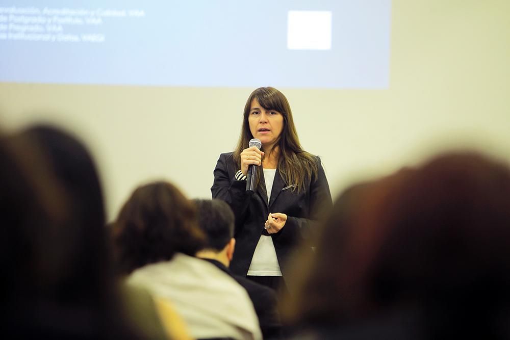 Se ve de frente, parada y presentando con un micrófono, a Cecilia Dooner, subdirectora de Aseguramiento de la Calidad de Pregrado de la U. de Chile.
