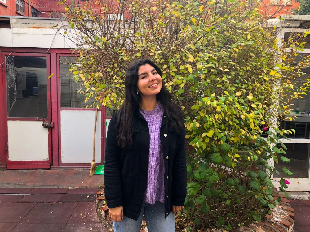 Constanza Ocampo, estudiante y tutora Uchile, sonríe de pie en el exterior, con un árbol detrás