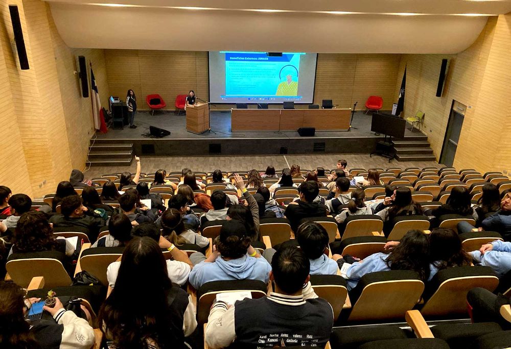 Auditorio lleno de estudiantes de educación media, asistiendo a una charla en la última feria de orientación a las y los postulantes Uchile. Adelante, monitores presentan un power point
