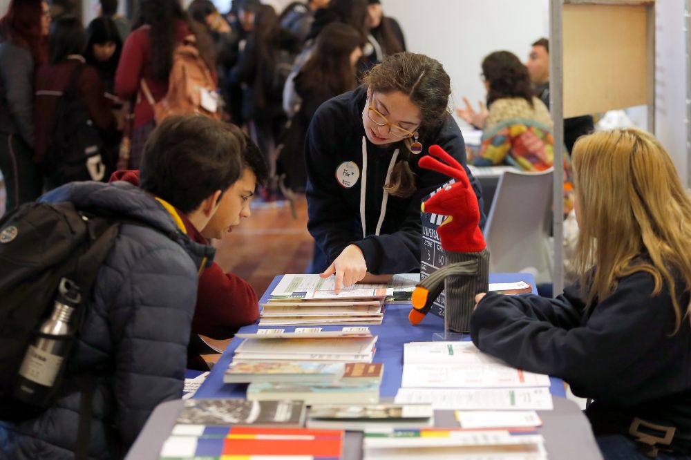 En un stand, se ve a una monitora explicándole mallas a dos estudiantes sentados.