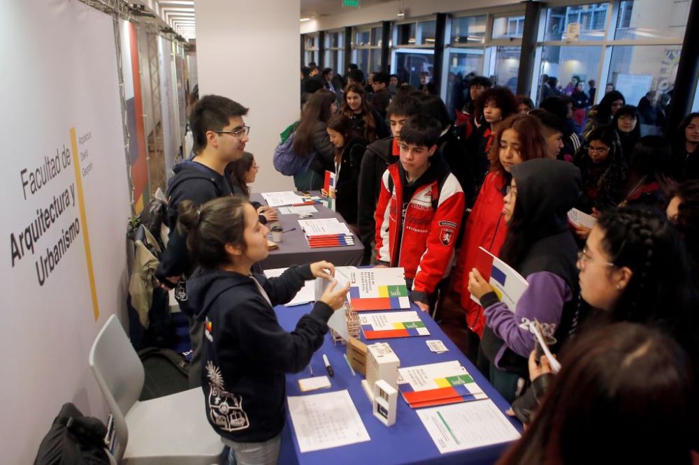 Se ve de frente a escolares escuchando a una monitora del stand de FAU, y mucha gente de fondo