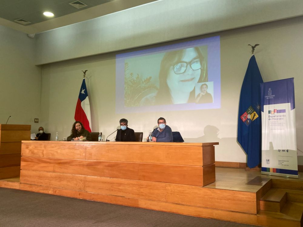 Tres acadèmicos en la testera y atrás en grande proyectada la imagen de una tercera académica que participa por videoconferencia