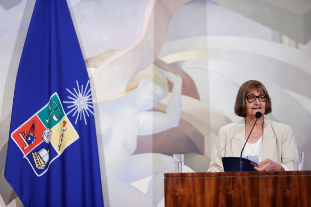 Rectora Rosa Devés en el estrado del Salón de Honor, hablando, con la bandera de la Universidad de Chile de fondo