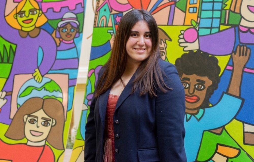 Medio plano de Charlotte Milling, sonriendo a la cámara y con un mural colorido con niños y niñas de fondo.