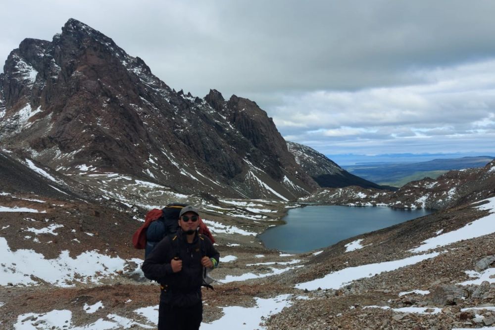 Medio plano de Matías Cuevas, con mochila, en en el circuito Dientes de Navarino, en medio de cimas nevadas y con una laguna detrás.