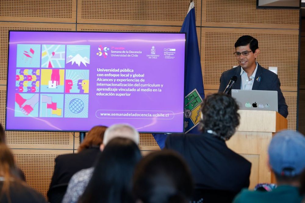Dr. Gerardo Blanco, hablando en la testera del auditorio. Al frente se ven las cabezas del público y al lado del profesor, una pantalla con la identidad gráfica de la Semana de la Docencia