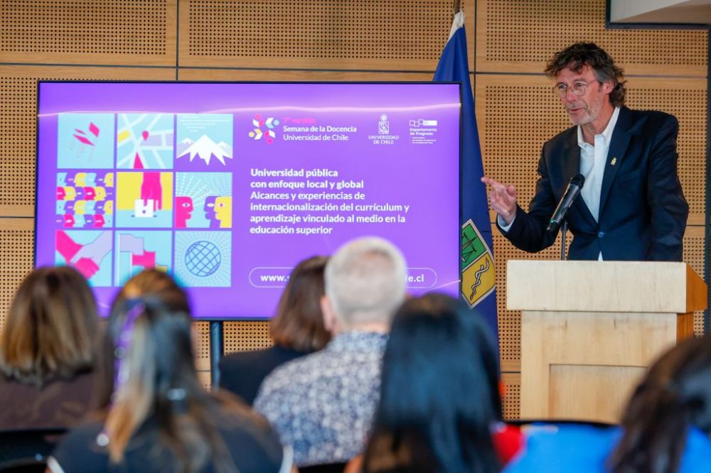 Vicerrector Pastenes, hablando en la testera del auditorio. Al frente se ven las cabezas del público y al lado del vicerrector, una pantalla con la identidad gráfica de la Semana de la Docencia