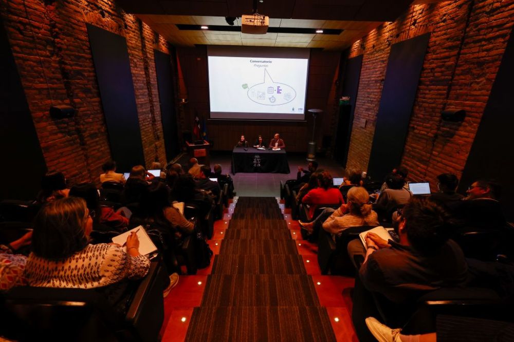Auditorio Facultad de Arquitectura y Urbanismo desde atrás. Se ve a las personas sentadas y adelante una mesa en que conversan las profesoras Rubliar y Armanet, además del profesor Olea.
