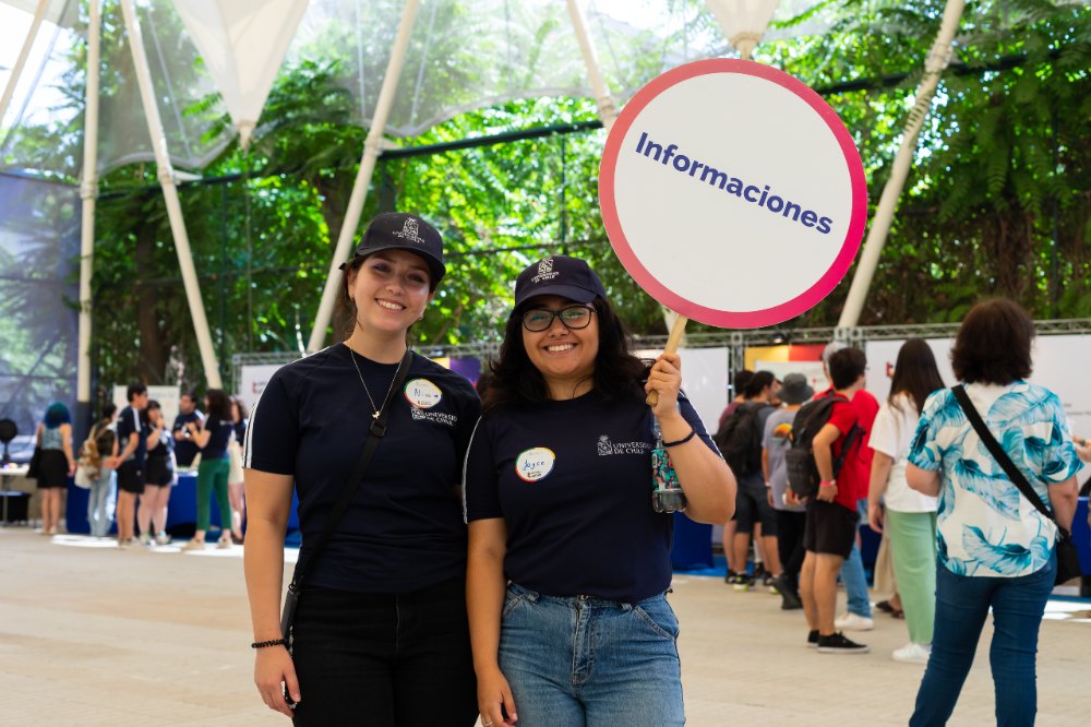 Dos monitoras posando a la cámara en una Semana de las y los Postulantes, con un cartel de informaciones en la mana y de fondo gente visitando el evento.