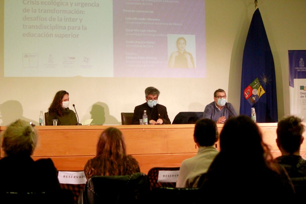 Tres académicos en la testera, observados por el público que se ve sentado de espaldas.