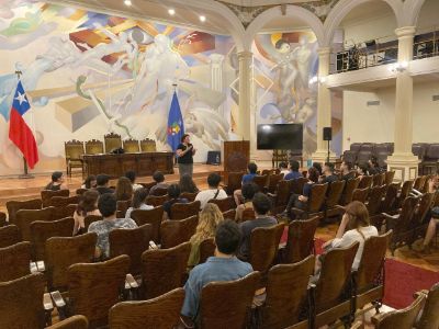 Profesora Ximena Póo se ve parada, de frente, en el Salón de Honor de Casa Central, dictando una clase. Estudiantes sentadas y sentados se ven por atrás.