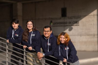 Cuatro estudiantes, dos mujeres y dos hombres, con el polerón de admisión uchile, posando para la cámara, apoyados en una baranda.