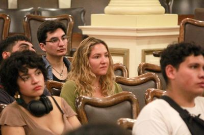 Medio plano de estudiantes sentadas y sentados en el Salón de Casa Central, poniendo atención.