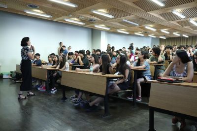 Decenas de estudiantes en un aulario, escuchando a profesora que está hablando. 