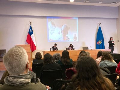 Vista hacia la testera de la Sala Eloísa Díaz de la Casa Central de la Universidad de Chile. Se ven las cabezas de personas mirando adelante a Theresa Lillis en la pantalla y a Federico Navarro y Leonor Armanet en la testera. Además, la intérprete de lengua de señas y las banderas de Chile y la U. de Chile