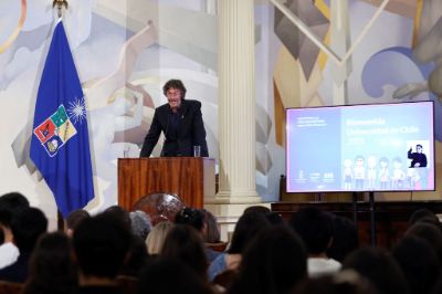 Vicerrector Claudio Pastenes, frente al público del Saló de Honor, a un lado la bandera de la U. de Chile y al otro la pantalla con banner de la bienvenida e interpretación de señas