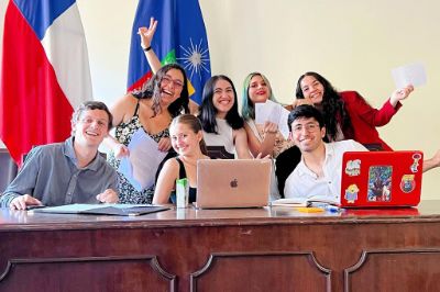 Grupo de siete estudiantes, posando con una sonrisa, con dos computadores al frente y las banderas de Chile y la Universidad de Chile, detrás