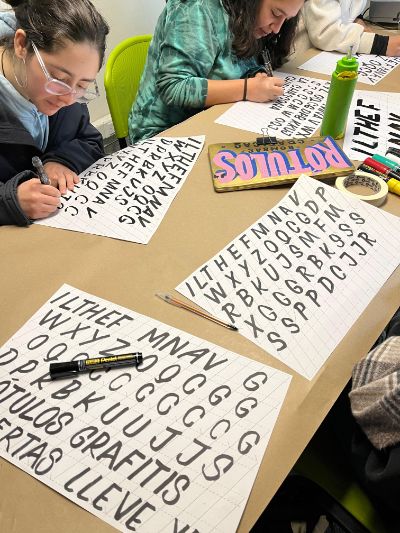 La imagen muestra a asistentes del taller de Rótulos y letreros a pincel dibujando letras.